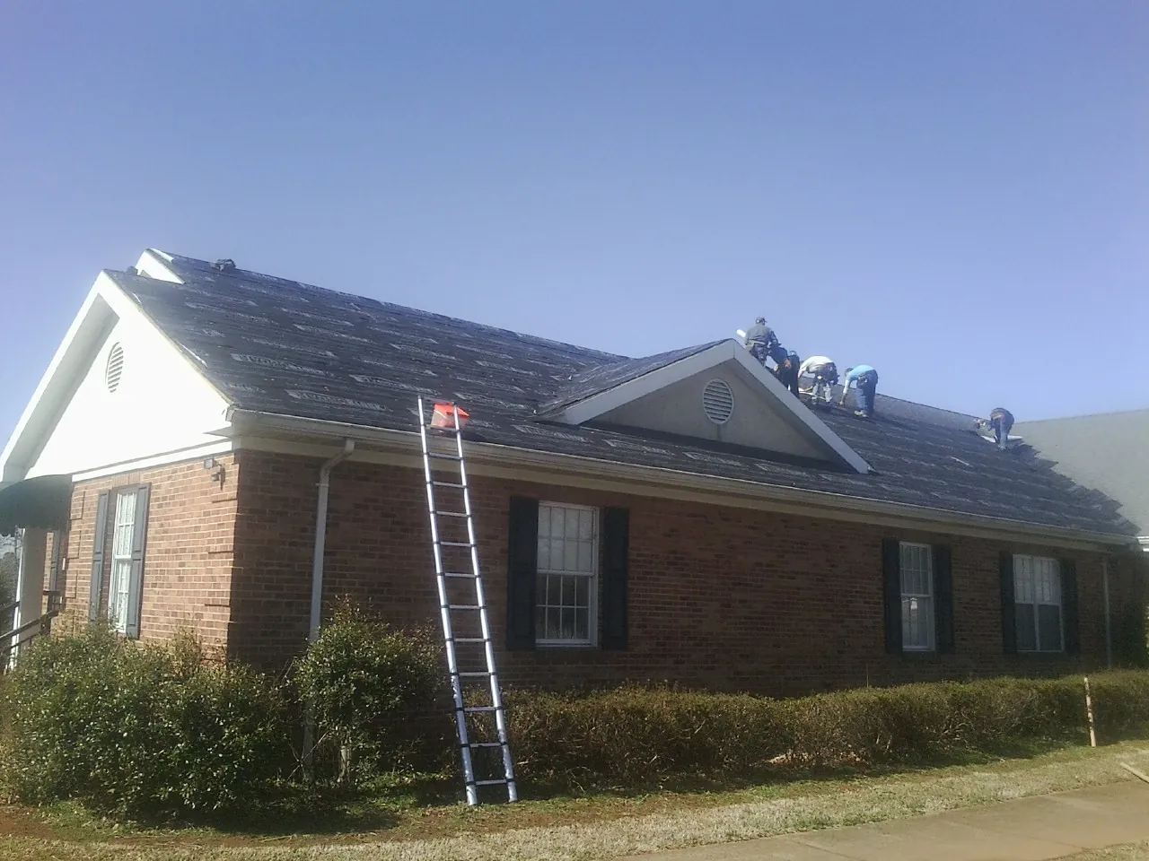 Crew tearing shingles off a brick ranch roof with a ladder set — Emergency Roof Repair work by our crew in Felda
