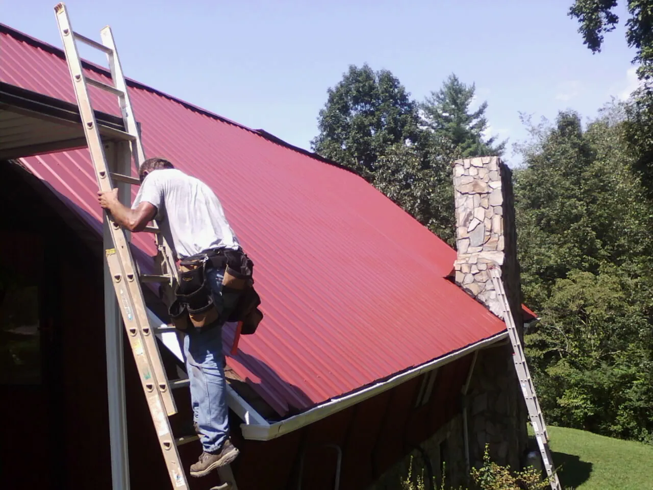 Roofer on a ladder beside a red metal roof and stone chimney — Soffit & Fascia Repair by Roofing Brothers in Felda